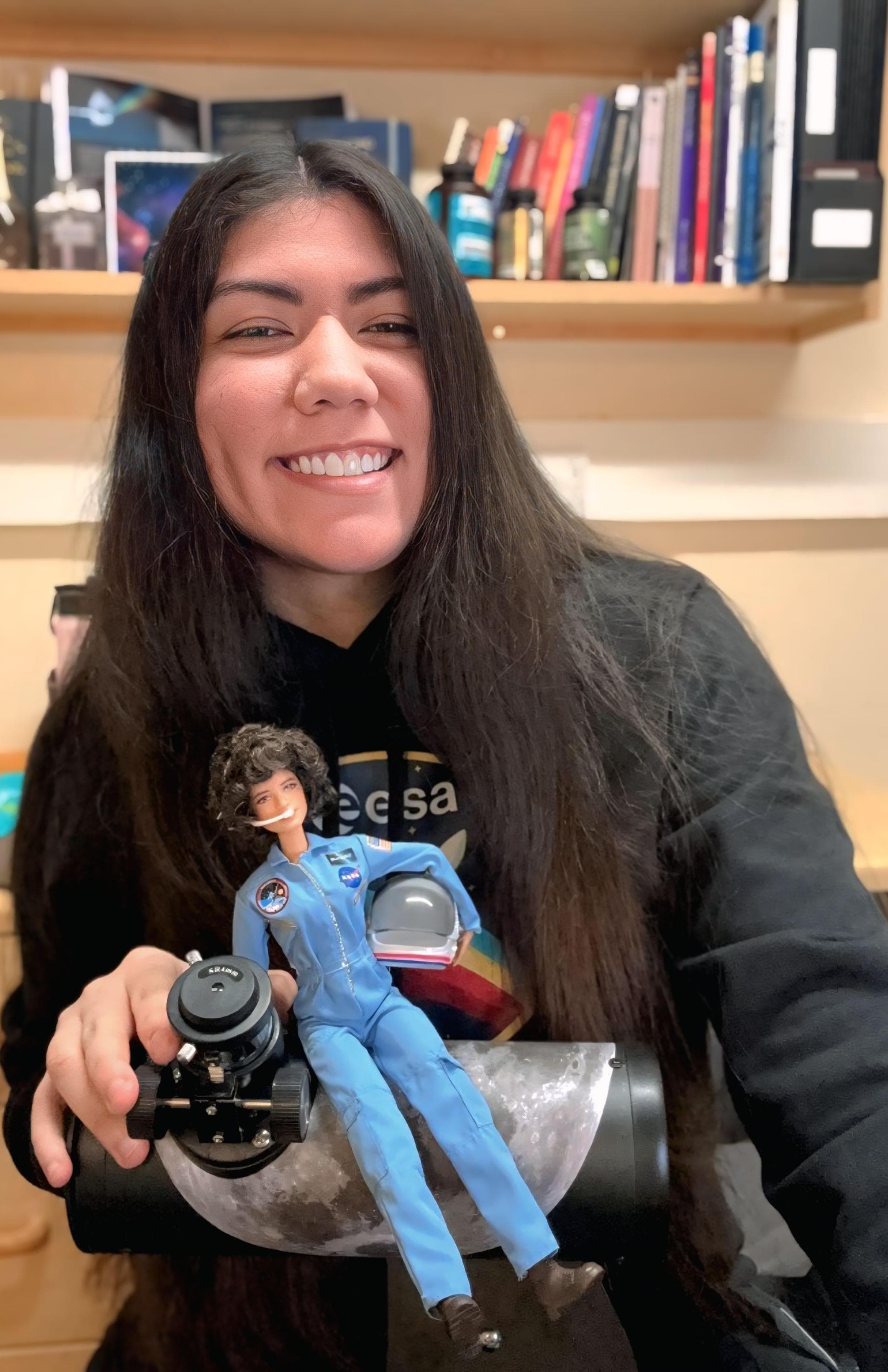 Carol Jiménez sonriendo en su escritorio, con una muñeca astronauta y un modelo de cohete en las manos