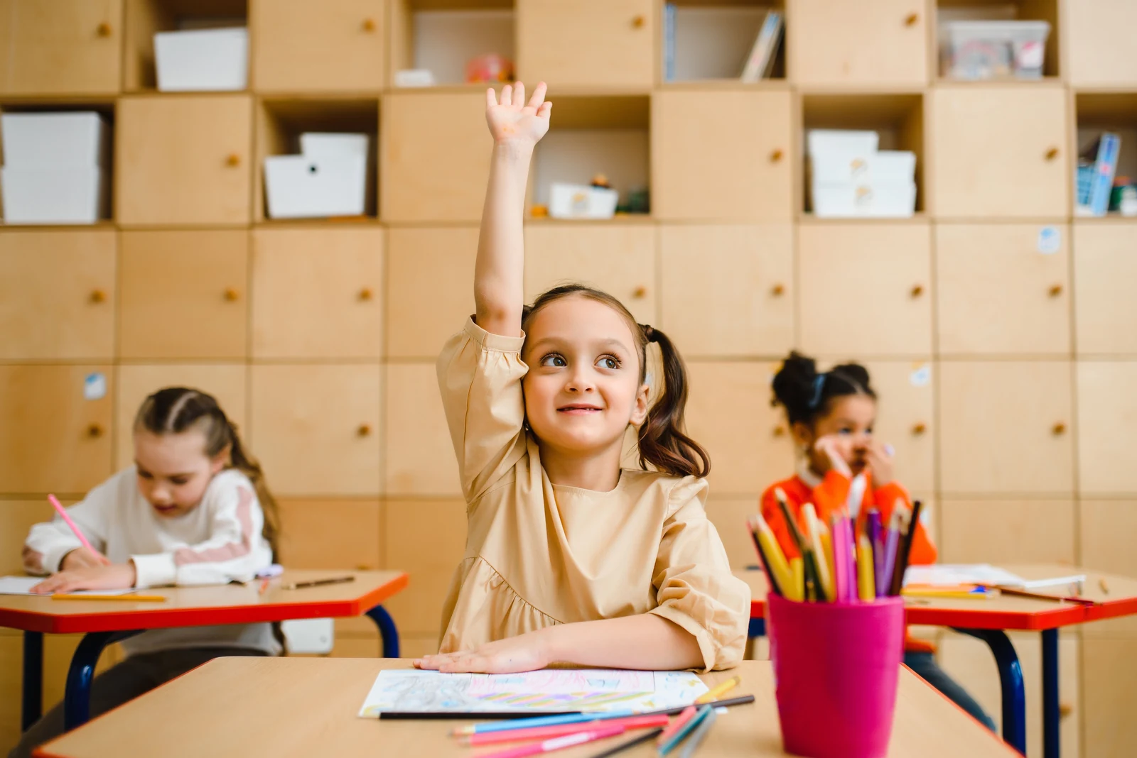 Una niña levanta la mano para participar mientras sus compañeras trabajan en clase